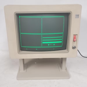 May include: A beige IBM computer monitor with a square screen displaying green text and grid lines. The monitor features a power switch on the right side, a key lock below the switch, and a sturdy base. The screen shows a green interface.