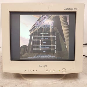 May include: A beige CRT computer monitor with the brand name SAMPO and AlphaScan 511 printed on the front bezel. The screen displays the main menu of the Age of Empires expansion game, featuring a classical architectural background and menu buttons.