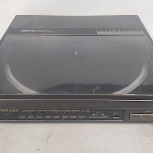Może przedstawiać: Czarny gramofon automatyczny Technics SL-Q3 Quartz Direct Drive. Gramofon ma przezroczystą, zakrzywioną osłonę przeciwpyłową i srebrny talerz. Przedni panel posiada przyciski sterowania i logo Technics.