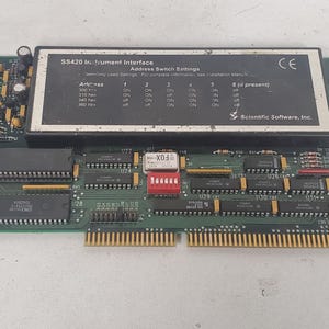 Op de afbeelding: Een groene printplaat met gouden connectoren, gemarkeerd met "SS420 Intrument Interface" en "Scientific Software, Inc." Beschikt over een zwart paneel met adres switch instellingen en een rode schakelaar. De kaart heeft diverse chips en een CE-markering.