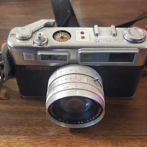 以下が含まれることがあります： シルバーとブラックのボディとYashinon-DX 1:1.7レンズを備えた、ヴィンテージのYashica Electro 35カメラ。 カメラには黒いストラップが付いており、木製の表面に置かれています。 レンズには数字付きのフォーカスリングがあります。