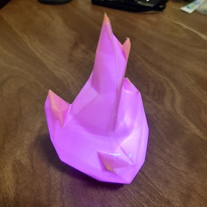 May include: A faceted, pink, crystal-shaped object with orange highlights. The object has a geometric design with sharp angles and a pointed top. The crystal is sitting on a wooden surface.