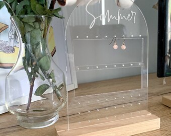 May include: A clear acrylic earring holder with a wooden base. The holder has a rounded arch at the top with the word "Summer" written in white. The holder has multiple holes for earrings and a pair of pink earrings are displayed.