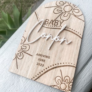 May include: A wooden arch-shaped sign with a floral design and the text "Baby" and "Compton" in white lettering. The sign also includes the text "Arriving June 2023".