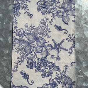 May include: A rectangular white paper napkin with a blue floral and marine life design. The design includes seahorses, starfish, seashells, and various flowers and coral. The napkin is folded and ready for use.