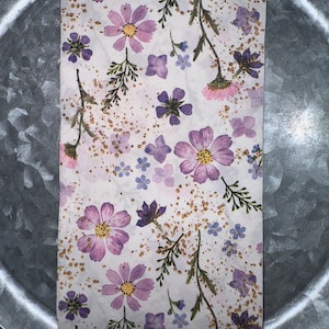 May include: A folded white paper napkin with a scalloped edge, adorned with purple and pink cosmos flowers, small blue flowers, and green sprigs. Gold flecks are scattered across the surface, creating a delicate, floral design.