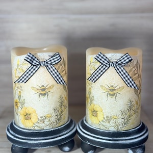 May include: Two decorative pillar candles with a bee and floral design. Each candle has a black and white checkered bow and sits on a black and white painted wooden base. The candles feature yellow flowers and bee illustrations.