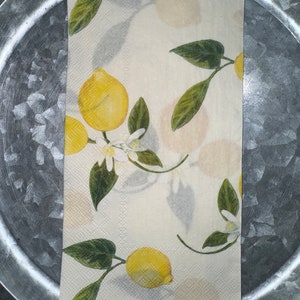 May include: A folded paper napkin with a lemon design. The napkin features yellow lemons, green leaves, and white blossoms on a cream-colored background. The napkin is placed on a silver metal tray.