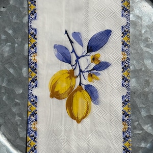May include: White paper napkin with a decorative border and a lemon branch design. The border features a blue and yellow pattern. The lemon branch has yellow lemons, blue leaves, and small yellow flowers.