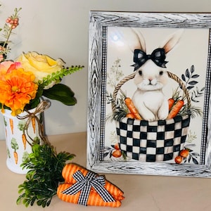 May include: A framed print features a white bunny with a black bow sitting in a basket with carrots. The basket has a black and white checkered pattern. A vase of flowers and a carrot-shaped decoration are also present.