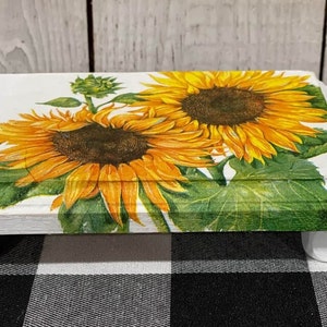 May include: A white, rectangular wooden riser with four rounded feet. The top surface is decorated with a vibrant image of two sunflowers with yellow petals and brown centers, surrounded by green leaves. The riser is suitable for home decor.