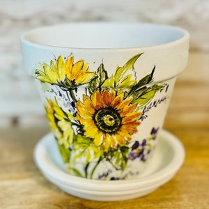 May include: White ceramic flower pot with a painted design of sunflowers and purple flowers. The pot has a matching saucer.