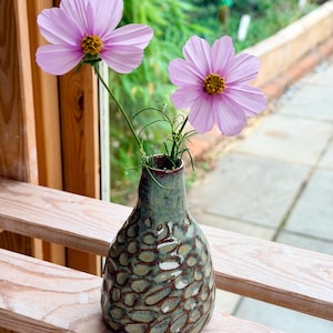 May include: A ceramic vase with a textured, mottled green and brown glaze featuring a repeating oval pattern. Two light pink cosmos flowers with yellow centres are arranged inside the narrow neck of the vase, extending upwards.