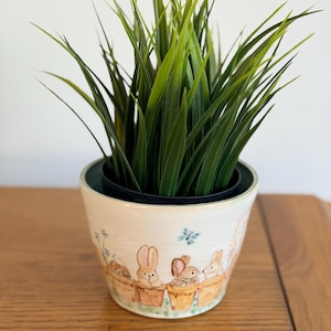 Könnte beinhalten: Ein dekorativer Keramik-Blumentopf mit einem bemalten Design von Hasen in Töpfen. Der Topf enthält eine leuchtend grüne Pflanze mit langen, schlanken Blättern. Der Topf steht auf einer Holzoberfläche.