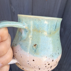 May include: A ceramic mug with a curved handle, featuring a two-tone glaze. The upper portion is a glossy light blue-green, while the lower section is a speckled beige. The surface has a smooth, reflective finish with visible dark brown speckles.