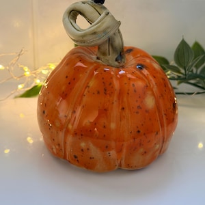 May include: A ceramic pumpkin sculpture featuring a glossy orange glaze with dark brown speckles throughout the body. The pumpkin has a prominent, coiled, light-coloured stem with a small dark blue accent at the very top, showcasing a smooth finish.