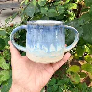 May include: A ceramic mug featuring two handles and a glaze that transitions from a deep blue at the rim to a lighter sky blue, ending in a solid cream-colored base. The surface has a smooth, glossy finish with visible drip patterns.