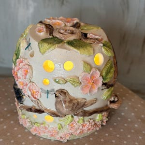 Puede incluir: Lámpara decorativa de cerámica con temática de la naturaleza. La lámpara presenta un pájaro, flores y un nido con huevos. La lámpara tiene una base de color crema con detalles en rosa y verde. La lámpara tiene varios agujeros que emiten un brillo amarillo cálido.