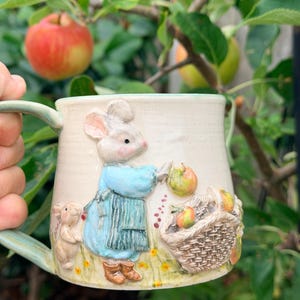 May include: A ceramic mug with a cream-colored base featuring a raised, three-dimensional relief of a mouse wearing a blue apron and brown boots. The mouse is picking apples near a woven basket filled with apples, set against a grassy field.