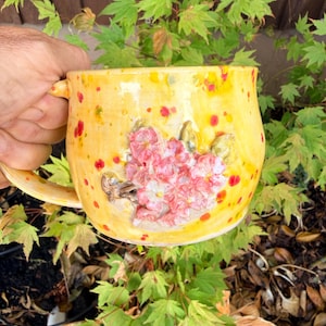 May include: A yellow ceramic mug featuring a speckled surface with red and orange dots. The mug is decorated with a raised, three-dimensional cluster of pink flowers and green leaves on the front, and includes a curved handle on the side.