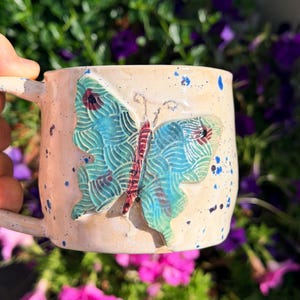 May include: A ceramic mug with a light beige base featuring blue and dark purple paint splatters. A raised, three-dimensional butterfly sculpture is attached to the front, decorated with teal, blue, and dark red patterns on its wings.