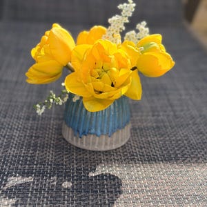Op de afbeelding: Een klein arrangement van gele tulpen en witte bloemen in een blauw en grijze keramische vaas. De vaas heeft een textuur met ribbels. De bloemen staan in volle bloei.