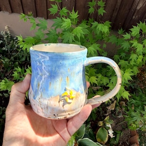 May include: A ceramic mug with a glossy finish featuring a blue and white gradient glaze. The mug has a curved handle and a small, three-dimensional yellow and white flower-shaped embellishment attached to the lower front exterior surface.