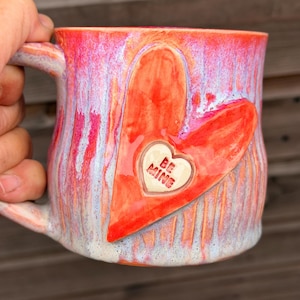 May include: A ceramic mug with a glossy glaze featuring vertical streaks of pink, orange, and white. A large, raised red heart is applied to the front, containing a smaller white heart-shaped emblem with the embossed text BE MINE.