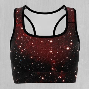 Op de afbeelding: Een zwarte sportbeha met een rode en zwarte galaxy print. De print heeft een donkere achtergrond met witte sterren en een rode nevel.