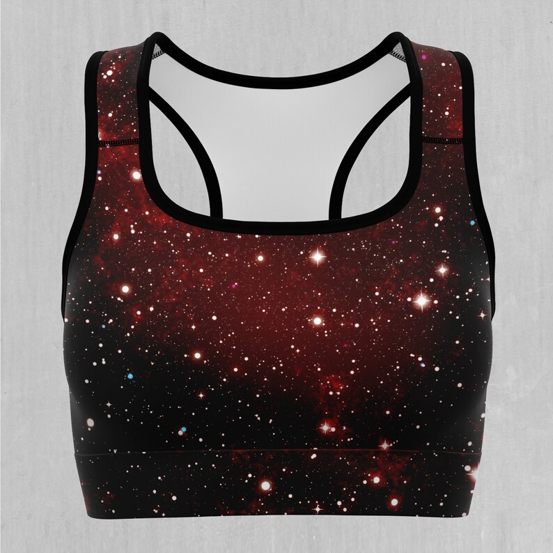 Op de afbeelding: Een zwarte sportbeha met een rode en zwarte galaxy print. De print heeft een donkere achtergrond met witte sterren en een rode nevel.