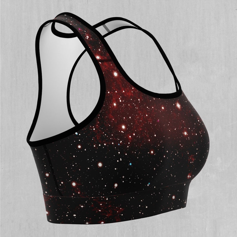 Op de afbeelding: Een zwarte sportbeha met een galaxy print met rode, witte en blauwe sterren. De beha heeft een racerback-stijl en is gemaakt van een rekbare, comfortabele stof.
