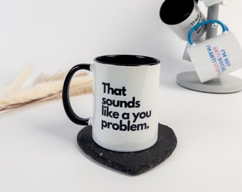 May include: White ceramic mug with a black interior and handle, featuring the text "That sounds like a you problem." The mug sits on a heart-shaped black coaster. Other mugs with humorous phrases are displayed on a mug tree.