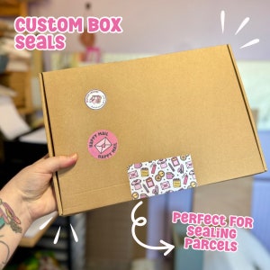May include: A brown cardboard box with pink and white custom box seals. The seals feature the text "Happy Mail" and a variety of illustrations. The box is being held by a hand.