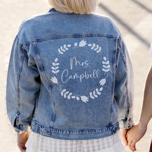 Peut inclure: Une veste en jean bleu clair avec un motif de couronne de fleurs blanches au dos. La couronne comprend le texte "Mrs. Campbell".