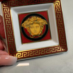 May include: Square ceramic trinket dish with a white interior and a red and gold border. The center features a Medusa head design on a black and red background. The dish is held by a hand.
