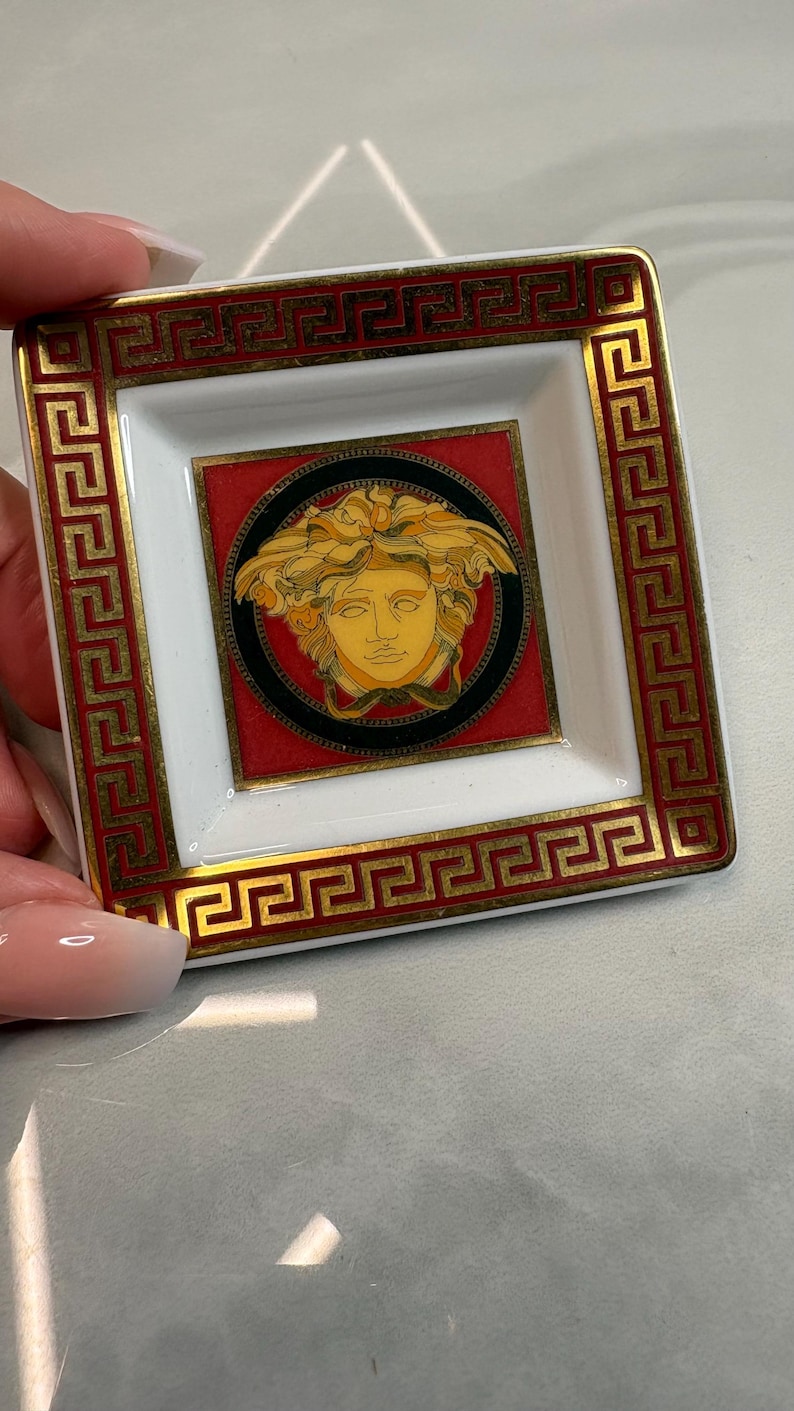 May include: Square ceramic trinket dish with a white interior and a red and gold border. The center features a Medusa head design on a black and red background. The dish is held by a hand.