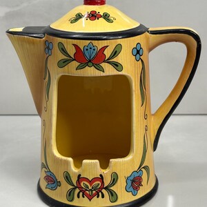May include: A yellow ceramic ashtray in the shape of a teapot, featuring a black handle and rim. The ashtray is decorated with floral designs in red, blue, and green. The lid has a black knob.