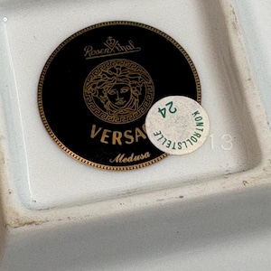May include: A white rectangular dish with a black and gold Versace Medusa logo. The logo is a circular design with the Versace name and Medusa's head. A small sticker with the number 24 is on the dish.