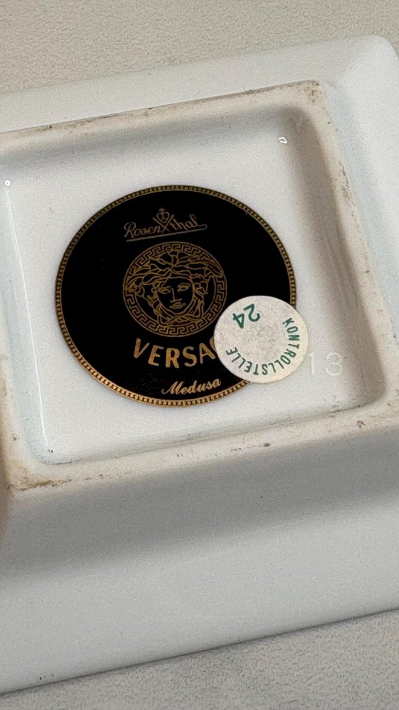 May include: A white rectangular dish with a black and gold Versace Medusa logo. The logo is a circular design with the Versace name and Medusa's head. A small sticker with the number 24 is on the dish.