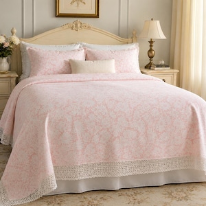 May include: A light pink bedspread featuring a white floral damask pattern and a wide, intricate white crochet lace border along the bottom edge. The set includes two matching pink floral pillow shams and a small, rectangular cream-colored throw pillow.