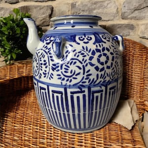 Pode incluir: Uma chaleira grande de cerâmica azul e branca com um bico e duas alças. A chaleira apresenta um padrão floral e geométrico detalhado. O fundo é uma cesta de vime tecida e uma parede de pedra.