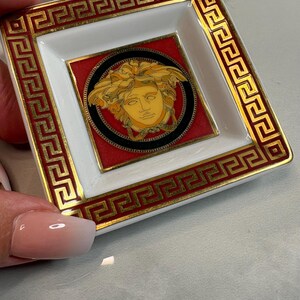 May include: Square, white and red decorative dish with a gold border and a central Medusa head design. The dish has a gold Greek key pattern around the edges. The Medusa head is gold and black.