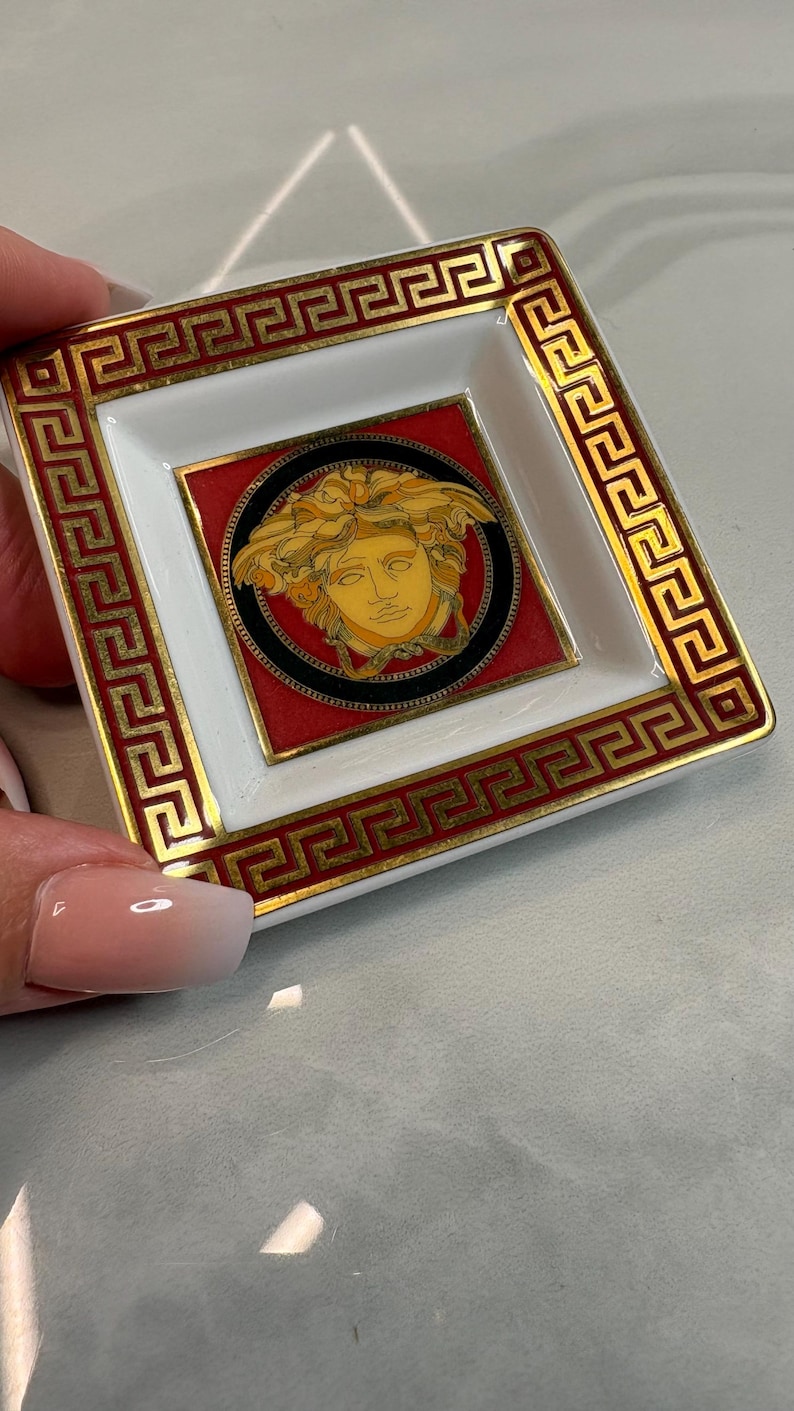 May include: Square, white and red decorative dish with a gold border and a central Medusa head design. The dish has a gold Greek key pattern around the edges. The Medusa head is gold and black.