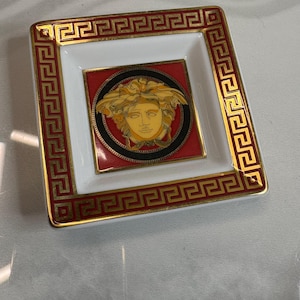 May include: Square decorative dish with a white and red interior, framed by a gold Greek key design. The center features a gold Medusa head on a red background, encircled by a black border. A luxury item.