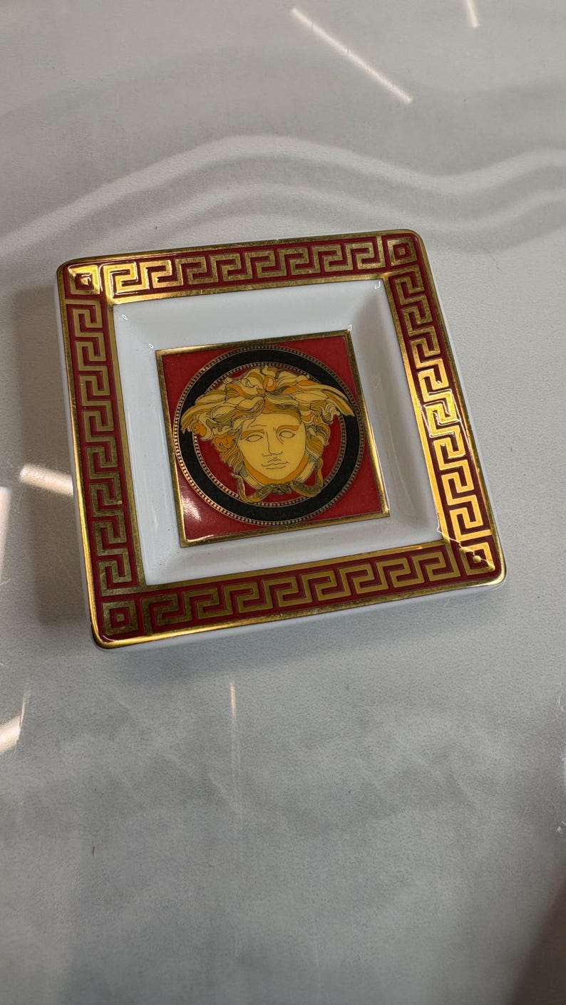 May include: Square decorative dish with a white and red interior, framed by a gold Greek key design. The center features a gold Medusa head on a red background, encircled by a black border. A luxury item.