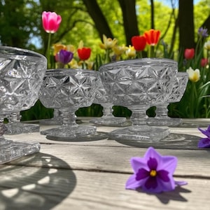 Puede incluir: Un juego de copas de postre de cristal transparente, de época, con diseños tallados en diamante. Las copas tienen una base de pedestal y un pie cuadrado. El fondo presenta tulipanes coloridos y una superficie de madera.