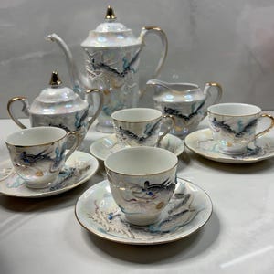 May include: A porcelain tea set with an iridescent finish featuring raised dragon motifs in blue, black, and gold. The set includes a teapot with a lid, a lidded sugar bowl, a creamer, and four teacups with matching saucers, all accented with gold trim.
