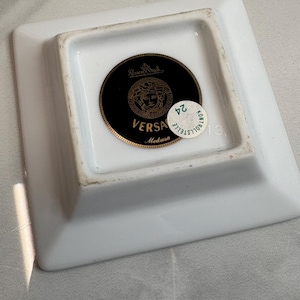 May include: Square white ceramic ashtray with a gold and black circular emblem featuring the Versace Medusa logo. The ashtray has a small, round, green and white sticker with the number 24.