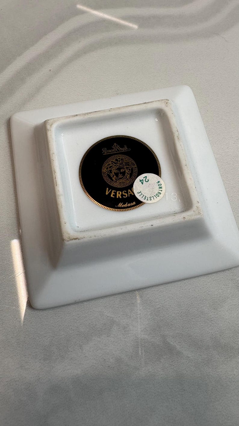 May include: Square white ceramic ashtray with a gold and black circular emblem featuring the Versace Medusa logo. The ashtray has a small, round, green and white sticker with the number 24.