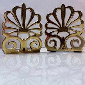 May include: A pair of polished brass bookends featuring an openwork design. Each piece displays a fan-shaped top section with radiating slats above a base decorated with symmetrical scrollwork patterns, creating a reflective metallic appearance.